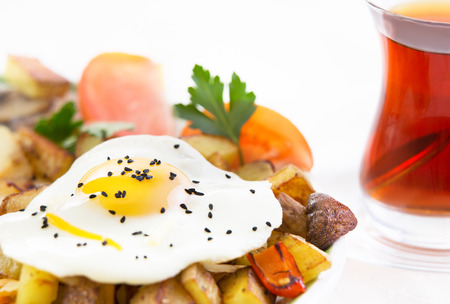 Sunny side up fried egg served on potatoes garnished with mushrooms and sliced fresh tomato accompanied by a glass of hot black Turkish tea on a white backgroundの写真素材