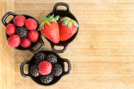 Overhead view of three ceramic ramekins full of healthy ripe fresh mixed berries including...