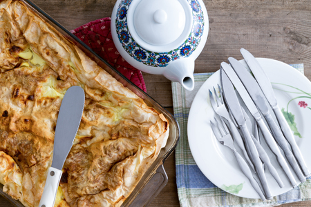 Overhead view of an oven dish with an uncut freshly baked Turkish borek ready for serving with a ceramic teapot and pile of plates and cutlery on a rustic wooden tableの写真素材