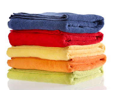Pile of neatly folded colorful cotton towels in red, blue, yellow, orange and green on a reflective white background with copyspace to the rightの写真素材