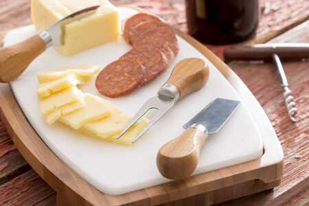 Harvati cheese with sliced spicy sausage served on a cheeseboard with cheese cutters alongside a bottle of red wine with a corkscrewの写真素材