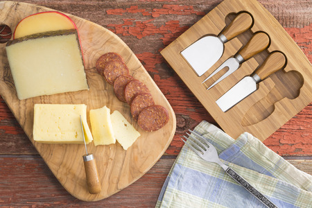 Overhead view of a cheeseboard with assorted cheeses including havarti, manchego and gouda served with sliced spicy sausage on a rustic grungy wooden table with a set of cheese cutters alongsideの写真素材