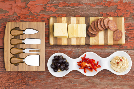 View from above of a selection of delicious savoury appetizers on a rustic wooden table with havarti cheese, spicy sausage, olives, red bell pepper and hummus served alongside a set of cheese cuttersの写真素材