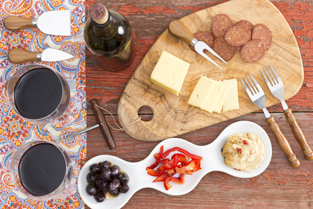 Overhead view of savory snacks on a rustic table with two glasses and a bottle of red wine and havarti cheese,spicy dried sausage,olives, red bell pepper and hummus with cheese cutters for servingの写真素材