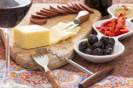 Havarti cheese and savory snacks including black olives, diced red bell pepper, hummus and spicy sausage served with a tall elegant glass of red wineの写真素材