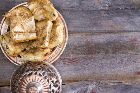 Portions of borek pastry served on a plate with a traditional silver metal dome set to one side on an old wooden rustic table with copyspaceの写真素材