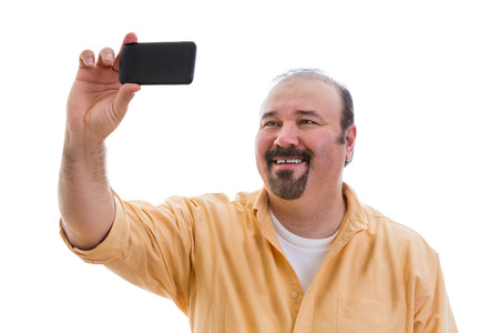 Happy man taking a self portrait or selfie on his mobile phone to send to his friends over the social media network holding the phone in his hand as he poses, isolated on whiteの写真素材