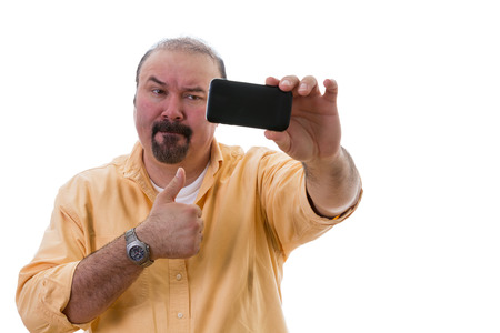 Middle-aged man with a goatee standing posing while taking a self portrait on his mobile as he gives a thumbs up of approval with a frown as though reluctant to do so, isolated on whiteの写真素材