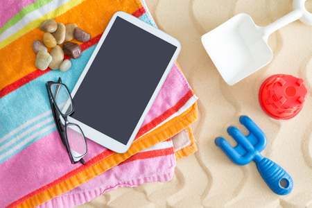 Tablet-pc lying on a colorful striped beach towel with glasses and waterworn stones on golden beach sand with a kids plastic toysの写真素材
