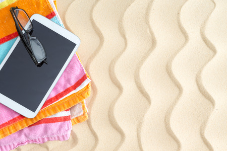 Tablet and glasses on a colorful beach towel lying on golden beach sand with a ridged wavy pattern in the hot summer sunshine for a relaxing day at the seasideの写真素材