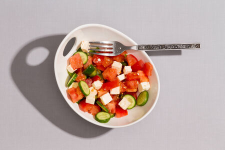 Bowl of fresh tasty Mediterranean salad made with fresh diced cucumber, feta cheese, tomato and green bell pepper ready for eating, overhead view on white with shadowの写真素材