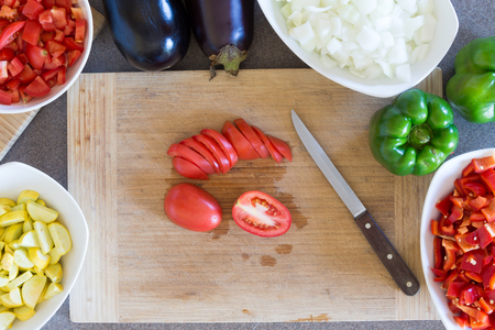 Prepping fresh vegetables for cooking in the kitchen with diced zucchini, tomato, white onion , green capsicum or bell pepper and eggplant arranged around a wooden chopping board with a knifeの写真素材