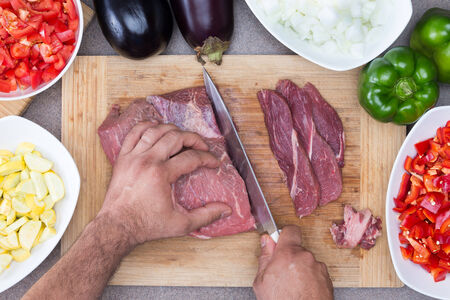 Hands of a man preparing meat and vegetables in a kitchen slicing lean meat on a wooden board with diced zucchini, tomato and onion and whole eggplant and green bell pepperの写真素材