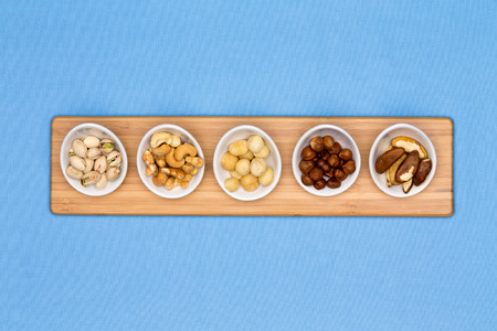 Taster dishes of assorted nuts containing cashews, pistachio, macadamia, hazel and brazil nuts arranged in a row on a wooden board centered on a blue background with copyspaceの写真素材