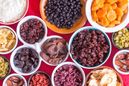 Colorful background of a variety of dried fruits in individual round dishes on red burlap with apple, goji berries, apricot, apple, banana and dates, view from aboveの写真素材