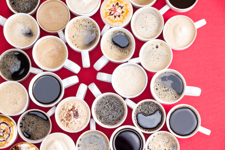 Coffee mecca with multiple assorted types and flavors of coffee in identical white mugs artistically arranged with converging handles in the centre on a red background, overhead viewの写真素材