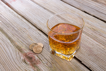 Tumbler of whiskey and tip of loose coins standing on an old rustic wooden bar counter with diagonal planks and copyspace, high angle viewの写真素材