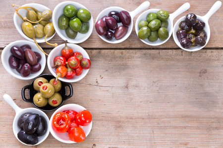 Border of assorted black, green and stuffed olives and green and red pimento peppers in small white ceramic dishes on a wooden counter with copyspaceの写真素材