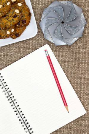 Open blank white notebook with lined pages and a pencil lying on a textured brown cloth with a plate of almond nut cookies and a a decorative round gift with a spiral pattern topの写真素材