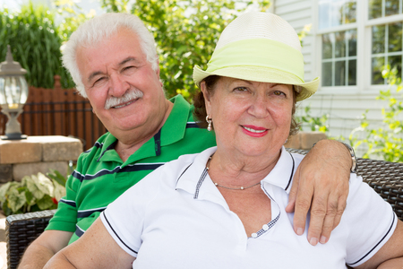 Attractive healthy happy senior couple sitting arm in arm on an outdoor summer patio in front of their home smiling warmly at the cameraの写真素材