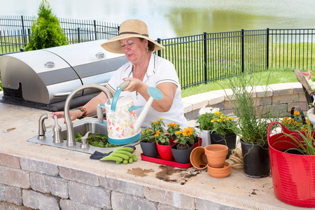 Senior lady filling a watering can on an outdoor patio as she prepares to water an array of potted plants lined up in her summer outdoor kitchenの写真素材