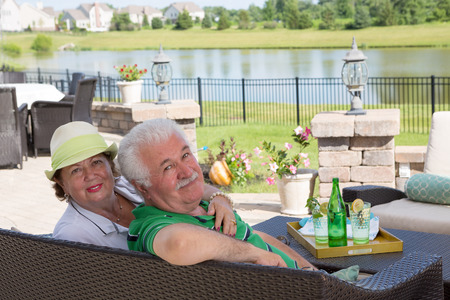 Affectionate elderly couple enjoy a relaxing day on the patio sitting close together on a wicker settee overlooking a scenic pond turning to look back at the camera with a smileの写真素材