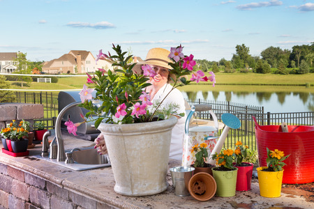 Happy grandmother working in her outdoor kitchen standing at the sink on the open-air patio in her sunhat surrounded by ornamental potted plants peeking through a gap at the camera with a lovely smileの写真素材