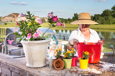 Elderly lady in a wide-brimmed straw sunhat tending to her potted plants on an outdoor patio overlooking a lake as she transplants and waters them in evening light with a delicate sunsetの写真素材
