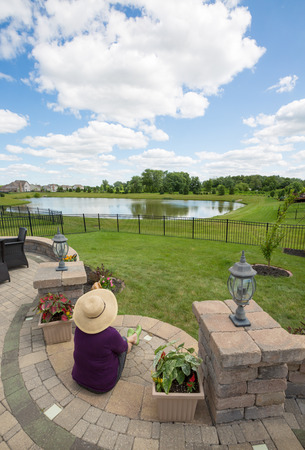 View from above of a woman in a straw sunhat sitting on her patio enjoying the view over green fields and a lake under a cloudy blue sky in spring sunshineの写真素材