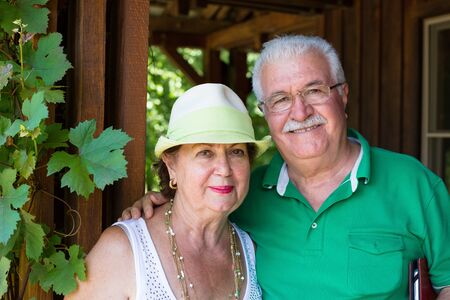 Happy trendy retired senior couple posing arm in arm on the porch of a wooden cabin alongside a green vine smiling happily at the camera with warm friendly smilesの写真素材