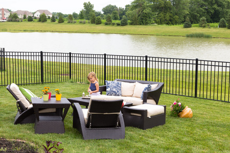 Cute little blond girl standing amongst comfortable modern design garden furniture with armchairs and tables arranged on a neat green lawn overlooking a tranquil pond and wrought iron railingの写真素材