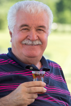 Smiling distinguished attractive senior man enjoying a glass of tea outdoors on a sunny summer afternoon smiling at the camera, focus to the glassの写真素材