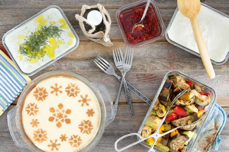 Delicious picnic lunch with grilled vegetables, tzatiki, yogurt with dill, pepper paste and a decorative milky semolina dessert , overhead view on a wooden tableの写真素材