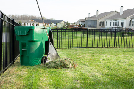 Raking lawn clippings on a neat upmarket suburban housing estate with a heap of grass cutting alongside a rake leaning on a green plastic bin for composting organic wasteの写真素材