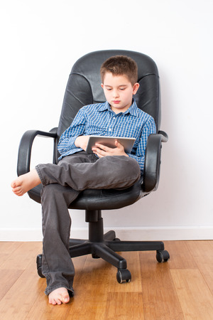 Busy Young Handsome Boy with Tablet Computer Sitting on a Black Office Chair with Legs Crossed and Bare Feet.の写真素材