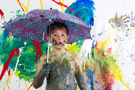 Cute funny shirtless little boy covered in fresh paint standing under an umbrella in front of his vibrant colorful contemporary artwork looking at the camera with an astonished lookの写真素材