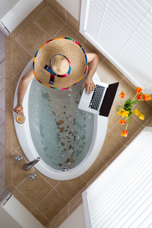 Man relaxing in a bath in a Mexican sombrero with a beer in one hand and using his laptop computer with the other, conceptual of a tourist on vacation in Mexico, overhead viewの写真素材