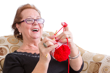 Happy elderly lady enjoying a joke laughing as she concentrates on her colorful festive red knitting while relaxing in a comfortable armchair, isolated on whiteの写真素材