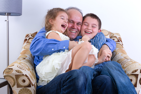 Middle-aged father with two laughing young kids, a girl and boy, on his lap sitting in an armchair enjoying a moment of fun and hilarity as a familyの写真素材