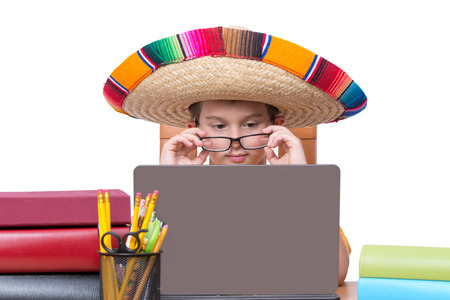Young Boy Wearing Eyeglasses and Colorful Mexican Sombrero Hat Looking Down at Laptop Computer Screen While Sitting at Desk Surrounded by Colorful Books and Binders and Writing Suppliesの写真素材