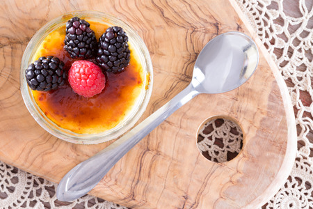 Scrumptious creme brulee topped with red and black raspberries from point of view angle with spoon over doily and wooden trayの写真素材