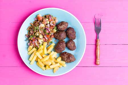 Plate of fresh homemade spicy traditional Turkish kofta meatballs served with a fresh lentil salad and fried potato chips served on a vibrant pink picnic table, overhead viewの写真素材