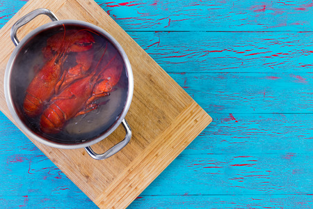 Two freshly boiled whole colorful red lobsters in a pot of water standing on a bamboo cutting board on a colorful blue crackle paint table, overhead view with copy spaceの写真素材