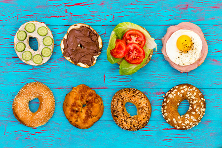 Four different flavored fresh sliced bagels with assorted filling of cucumbers, chocolate , salad, fried egg and ham, neatly arranged in a row on an exotic blue picnic table viewed from aboveの写真素材