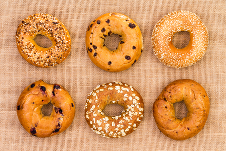 Six different tasty fresh ring-shaped bagels displayed in neat rows on burlap viewed from above