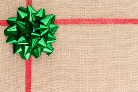 Christmas background on natural burlap textile with a decorative red ribbon and green foil bow with metallic sheen in the corner, large copy space for your seasonal wishesの写真素材