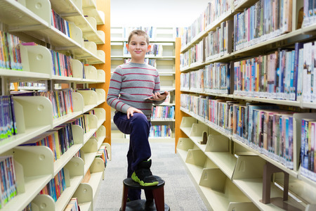 Happy boy in library surrounded by book shelves with foot on stoolの写真素材