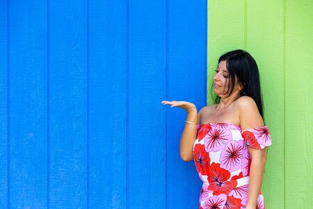 Young woman looking at her empty hand extended for product placement as she stands in a red floral summer dress in front of a blue and green timber wall with copy spaceの写真素材