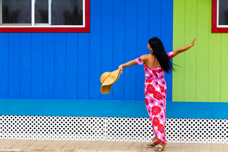Pretty Polynesian islander in a red floral dress and sunhat dancing in front of a colorful green and blue wooden wall in Hawaiiの写真素材