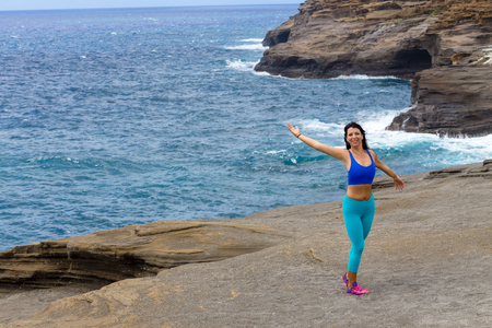 Carefree young woman exercising at Heavens Point, Oahu, Hawaii standing smiling with her back to the pacific Ocean and outstretched armsの写真素材